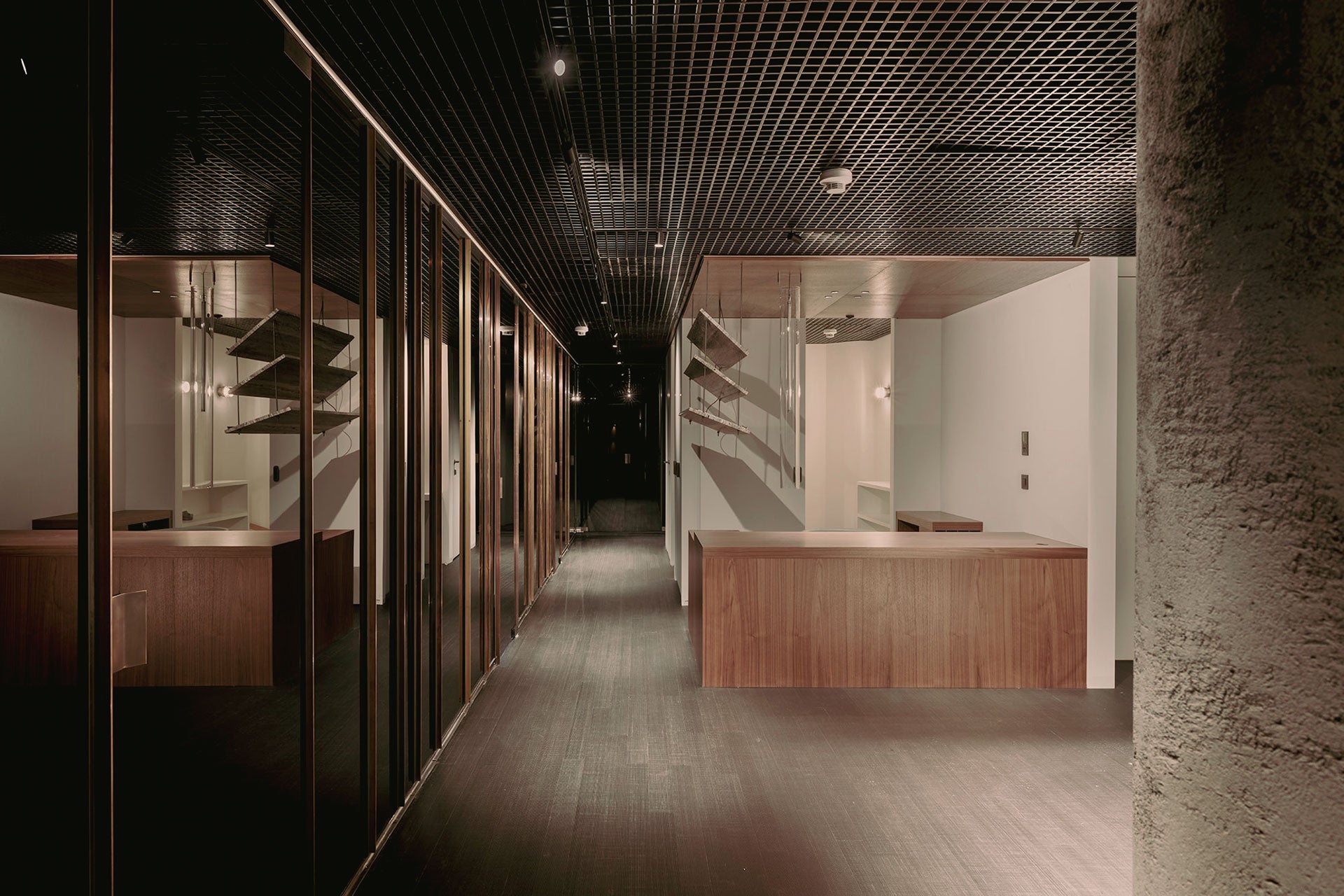 Classy hallway with dark wooden floors and a metal grid ceiling, featuring a wooden reception desk, travertine marble magazine shelf, and room facades with metal profiles and opaque glass, illuminated by ambient lighting.