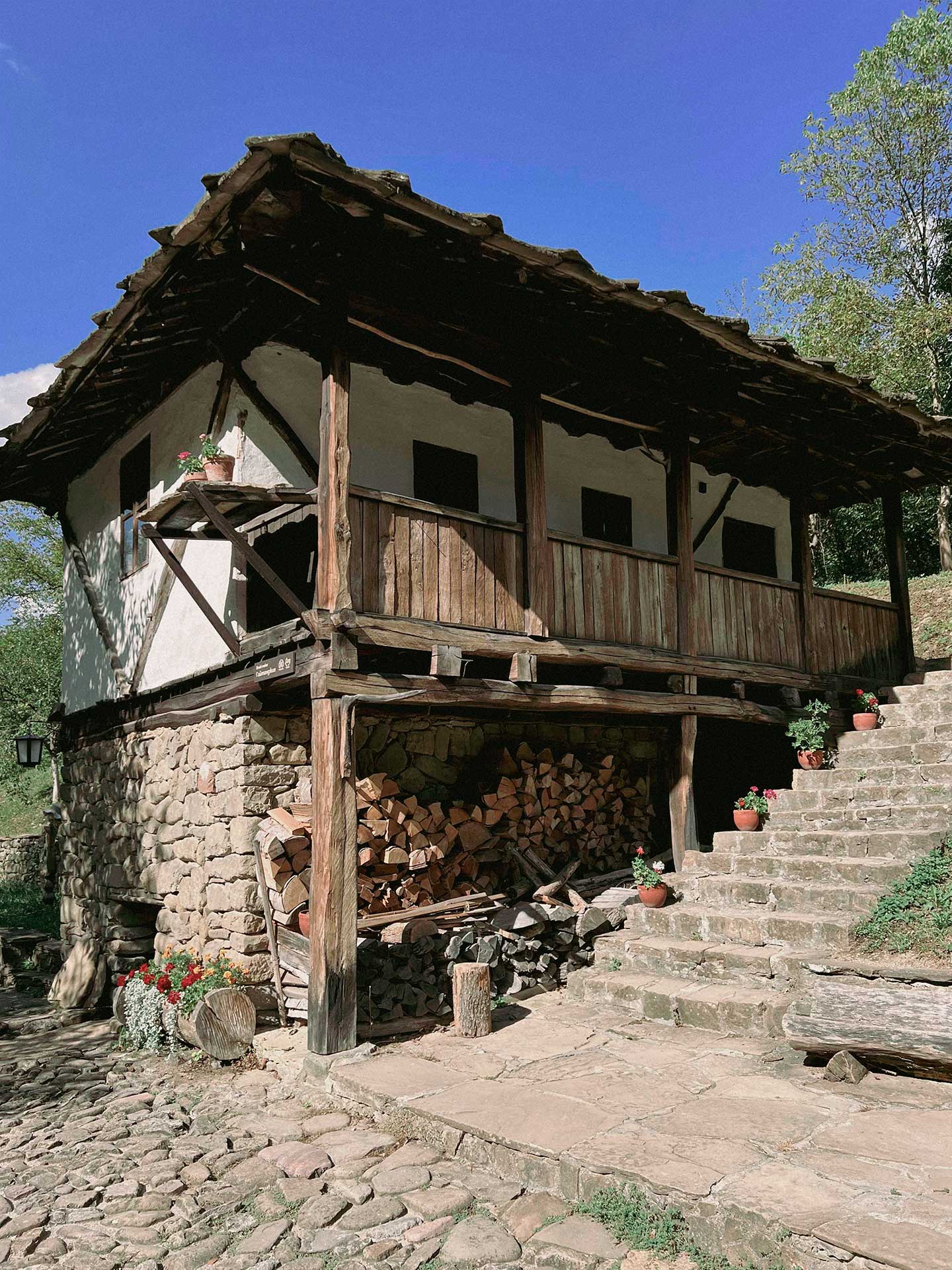 Traditional Balkan house in Bulgaria's Troyan region with white plaster walls, wooden construction roof, stone base, firewood stacked under the house, stone paving and steps, set against a clear blue sky.